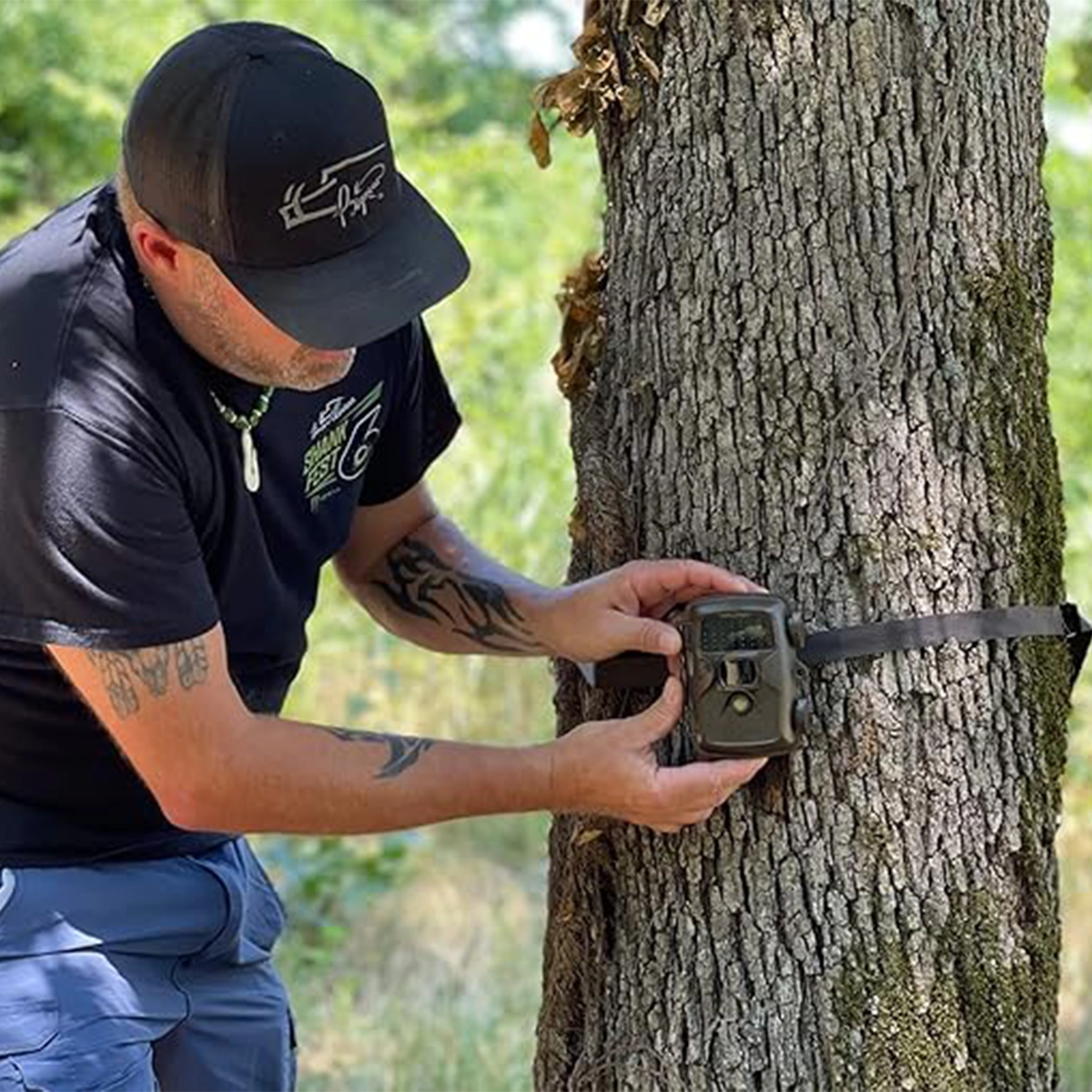 Covert MP30 Trail Camera 3 A person secures a Covert MP30 Trail Camera to a tree trunk with a strap in an outdoor, wooded area.