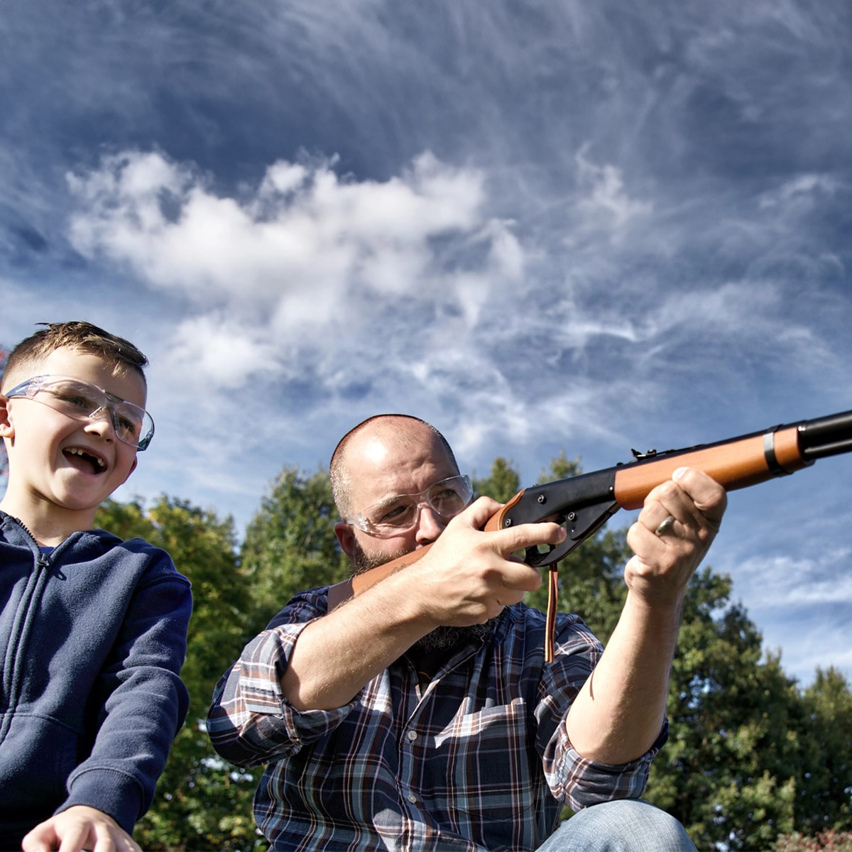 Daisy Red Ryder Spring BB Air Rifle Kit 3 An adult and a child, both in safety glasses, aim the Daisy Red Ryder Spring Air Rifle Kit outdoors under a blue sky with clouds, surrounded by green trees.
