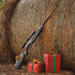 A rifle and an airgun rest against a large hay bale outdoors, with two wrapped Christmas gifts placed beside them.