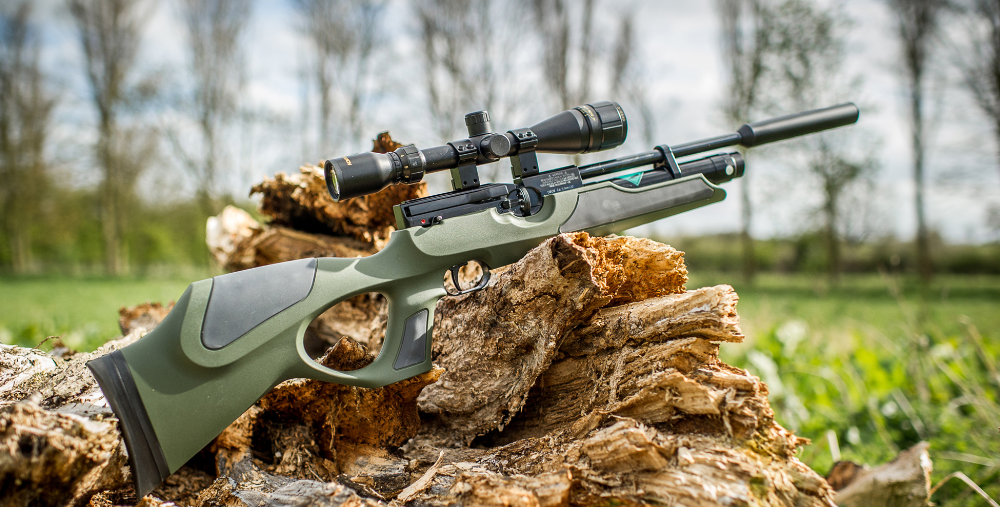A Beginners Guide To PCPs 1 A scoped air rifle with a green and black stock, perfect for beginners, rests on a pile of logs outdoors—much like how PCPs guide patients; trees and grass provide a natural backdrop.