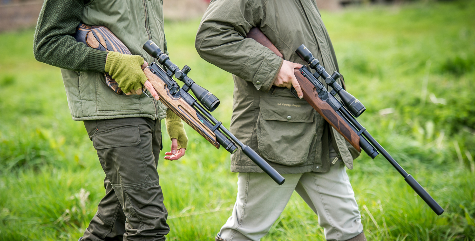 What & Where Can I Shoot? 1 Two people walk on grass, each holding a scoped rifle with a silencer, wearing outdoor jackets and gloves—perfect for those seeking new shooting locations or following a trusted shooting guide.