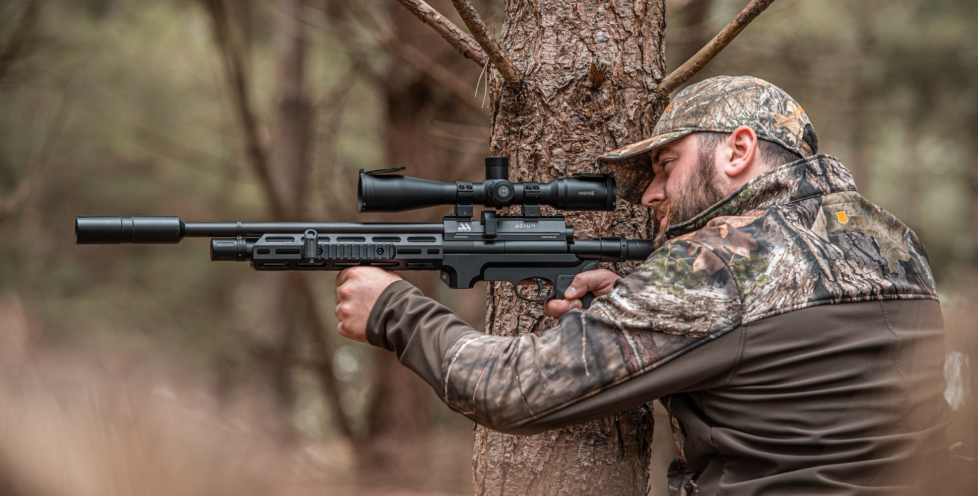A man in camouflage clothing aims a scoped rifle, using a tree for support in a wooded outdoor setting, focusing intently on proper rifle sighting.