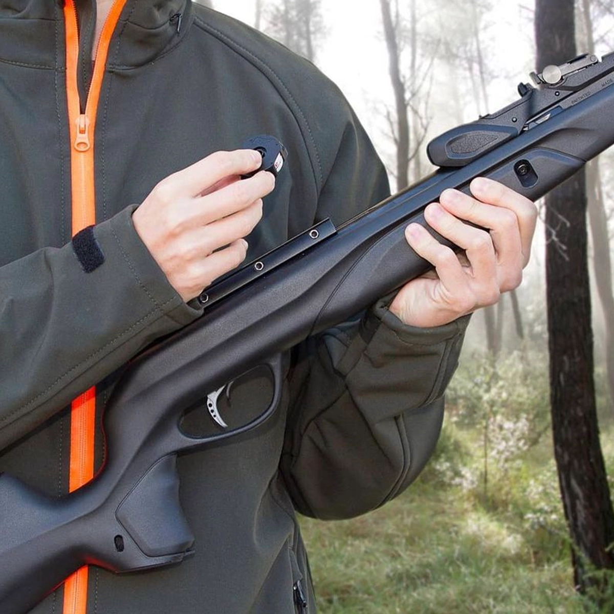 Gamo Swarm Speedster Gen 2 Multi Shot Spring Air Rifle With Scope 11 A person in an outdoor setting loads a pellet magazine into a Gamo Swarm Speedster Gen 2 Spring Air Rifle With Scope, while wearing a green jacket with an orange zipper.