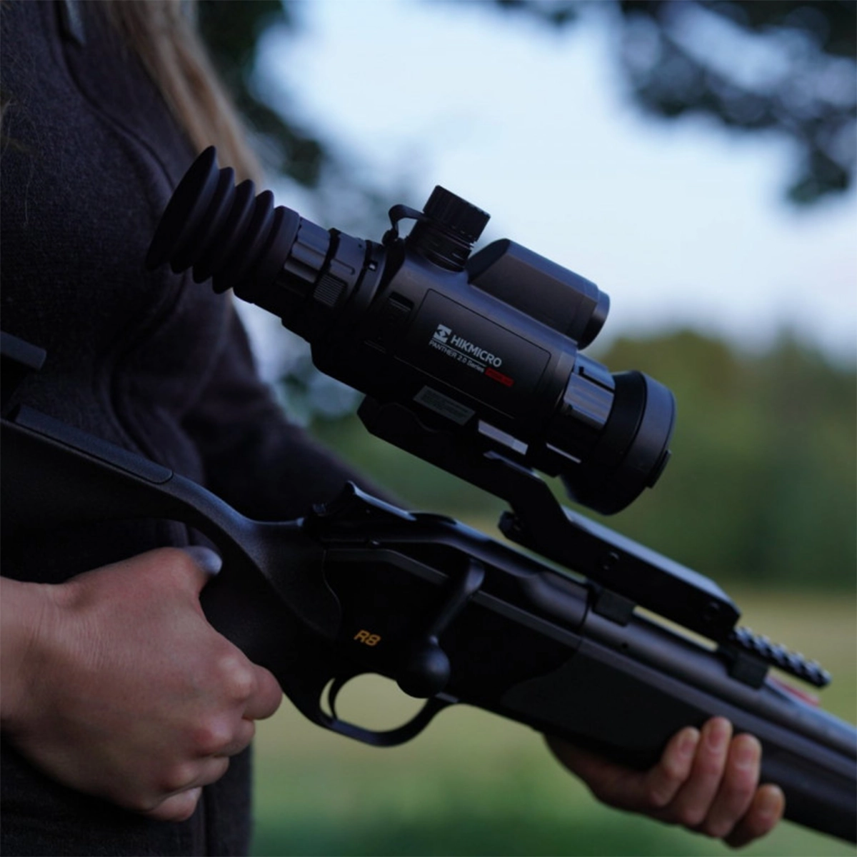 Hikmicro Panther 2.0 PQ50L Thermal Imaging LRF Rifle Scope 11 A person holds a rifle outfitted with the Hikmicro Panther 2.0 PQ50L Thermal Imaging LRF Rifle Scope outdoors, with blurred trees and sky in the background.
