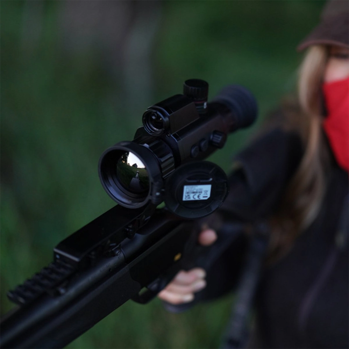 Hikmicro Panther 2.0 PQ50L Thermal Imaging LRF Rifle Scope 13 A person in a red face covering and cap holds a scoped rifle, highlighting the Hikmicro Panther 2.0 PQ50L Thermal Imaging LRF Rifle Scope and its advanced electronic features.