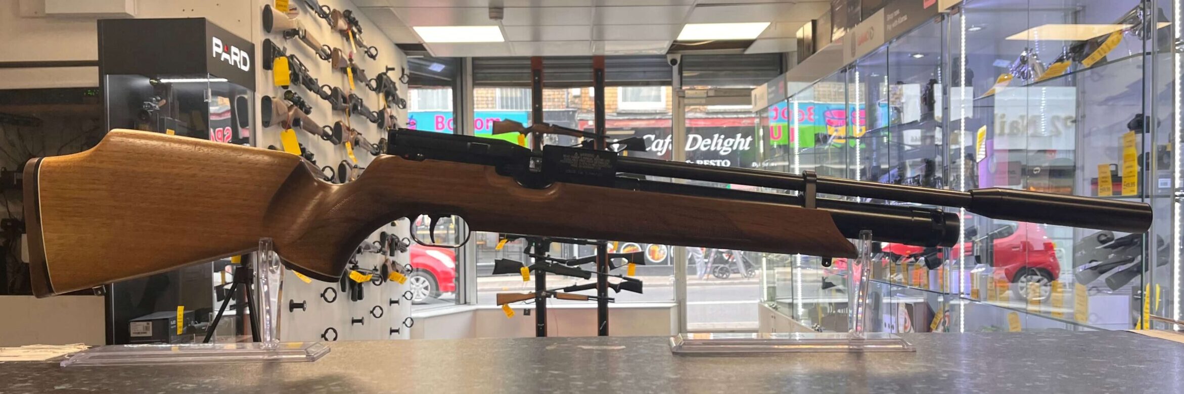 PRE-OWNED Weihrauch HW100 PCP Air Rifle Walnut .22 1 A PRE-OWNED Weihrauch HW100 PCP Air Rifle Walnut .22 is shown on a counter in a bright gun shop, with shelves and walls behind it filled with firearms and accessories.