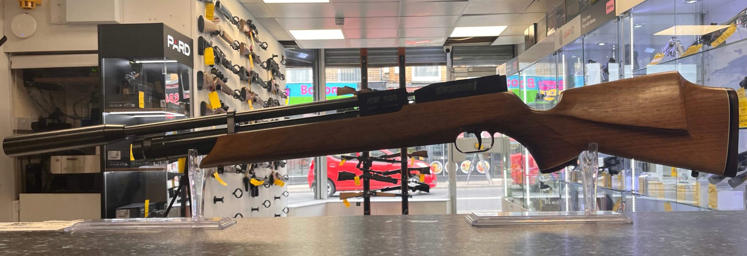 PRE-OWNED Weihrauch HW100 PCP Air Rifle Walnut .22 3 A PRE-OWNED Weihrauch HW100 PCP Air Rifle Walnut .22 is displayed on a counter in a well-lit shop, with various firearms and accessories visible on the shelves and walls behind it.