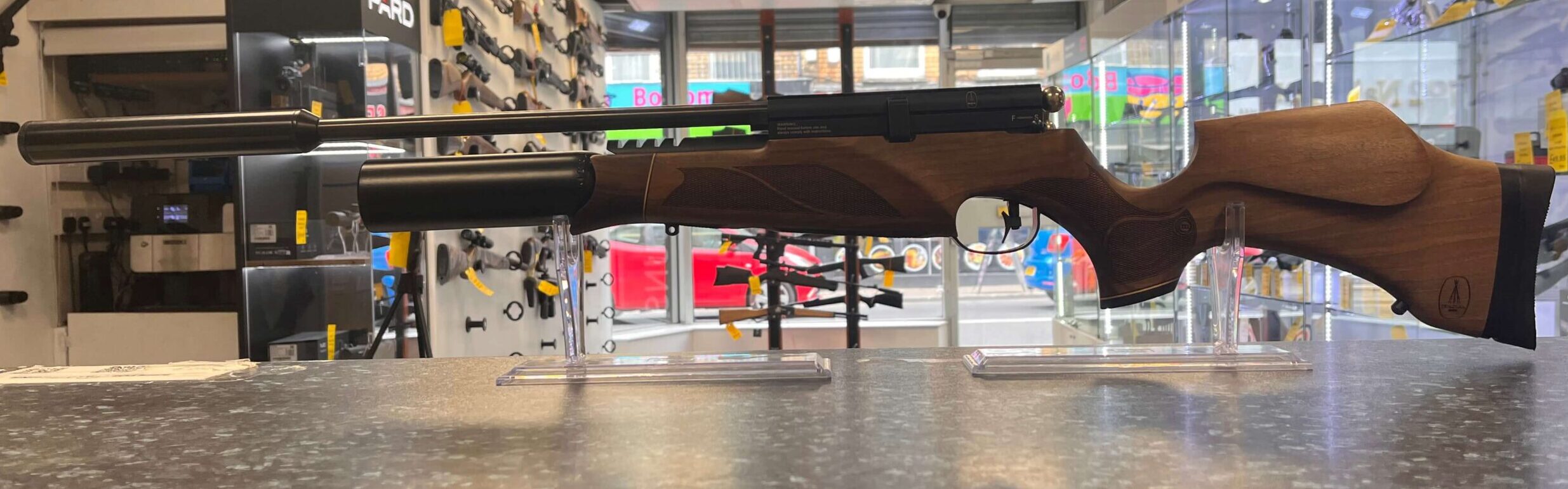 PRE-OWNED BSA R10 MKII PCP Air Rifle .22 3 A PRE-OWNED BSA R10 MKII PCP Air Rifle .22 with a wooden stock is displayed on a counter in a brightly lit gun shop, surrounded by various .22 accessories and equipment on shelves and walls in the background.