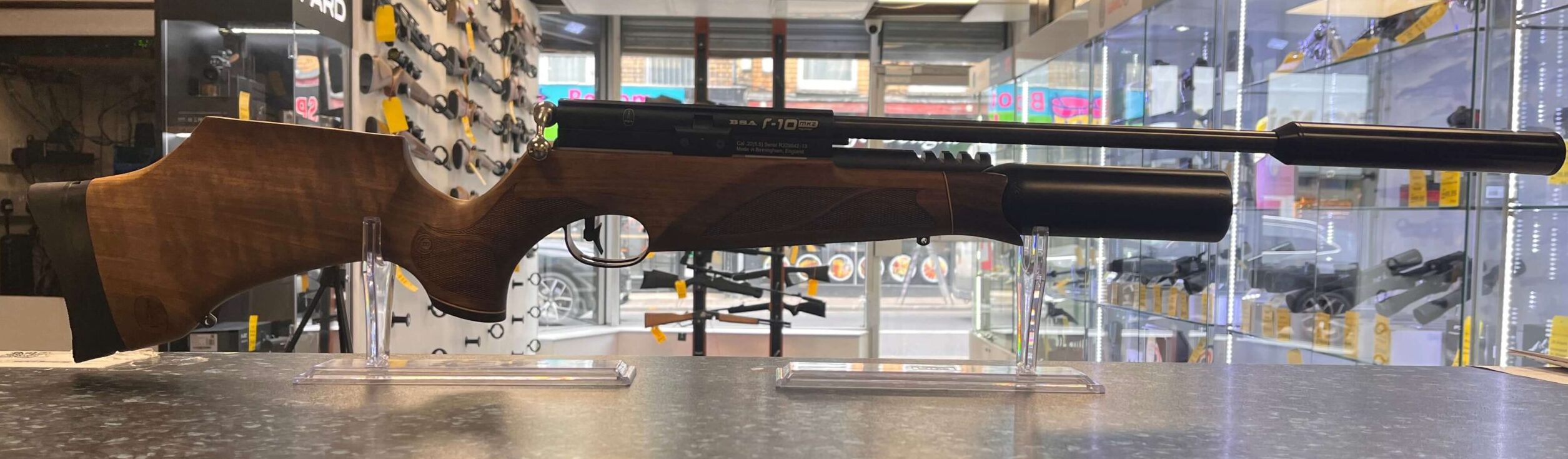 PRE-OWNED BSA R10 MKII PCP Air Rifle .22 1 A PRE-OWNED BSA R10 MKII PCP Air Rifle .22 with a wooden stock and black barrel is displayed on a grey countertop in a shop, surrounded by other firearms and accessories visible on shelves in the background.