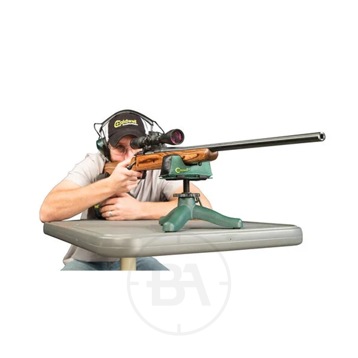 Caldwell Rock JR Shooting Rest 3 A person with ear protection aims a scoped rifle supported on a Caldwell Rock JR Shooting Rest atop a gray table.