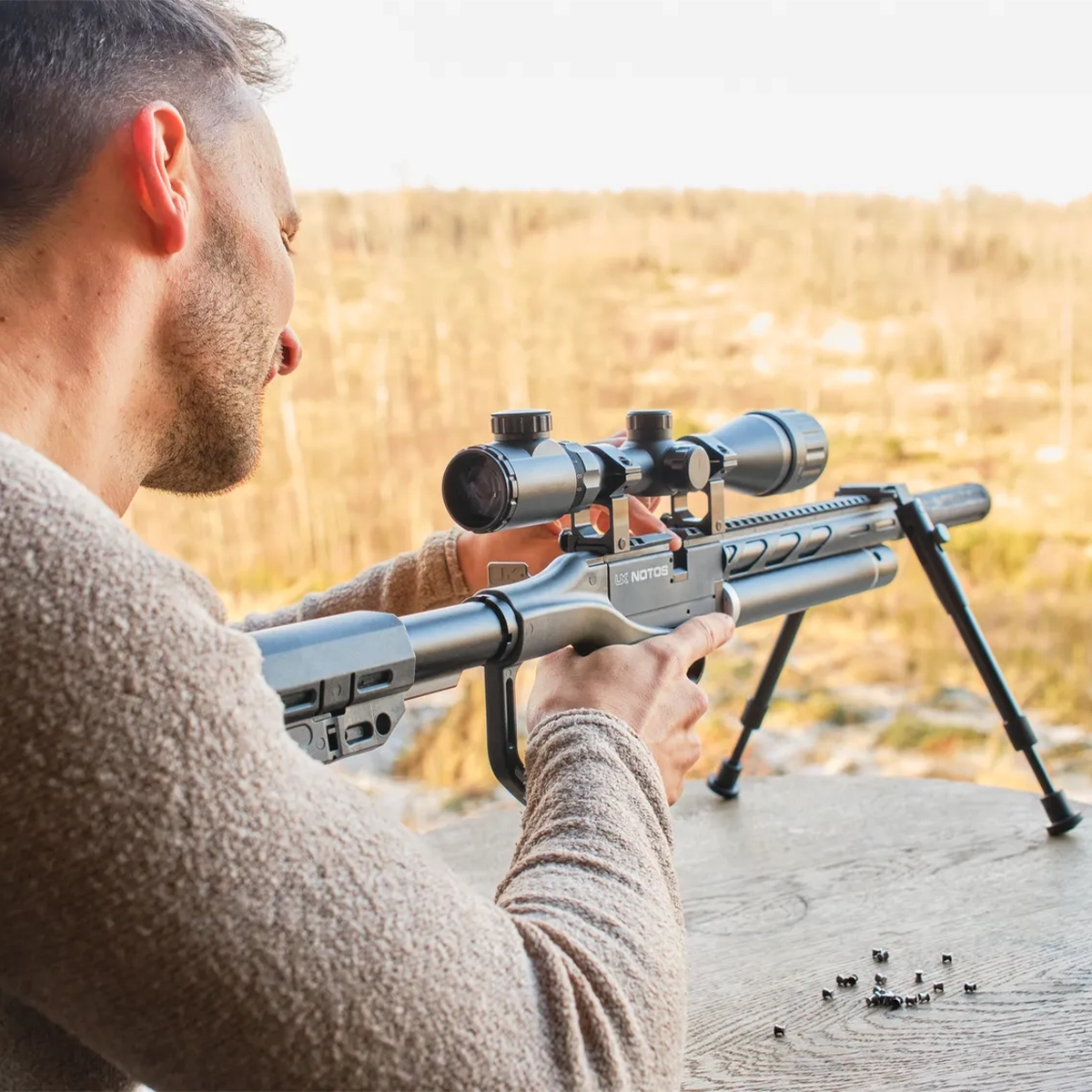 Umarex UX Notos PCP Air Rifle Kit With Scope & Bipod 7 A person aims an Umarex Notos PCP Air Rifle Kit with Scope & Bipod on a wooden surface outdoors, pellets scattered nearby, with a blurred landscape in the background.