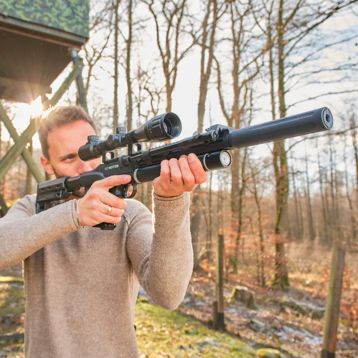 Umarex UX Notos PCP Air Rifle Kit With Scope & Bipod 9 Outdoors, a person aims a Umarex Notos PCP Air Rifle Kit with Scope & Bipod; trees and a wooden structure can be seen in the background.
