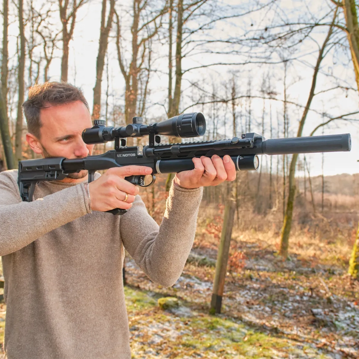 Umarex UX Notos PCP Air Rifle Kit With Scope & Bipod 11 A man in a beige sweater aims the Umarex Notos PCP Air Rifle Kit With Scope & Bipod outdoors in a wooded area during daylight.