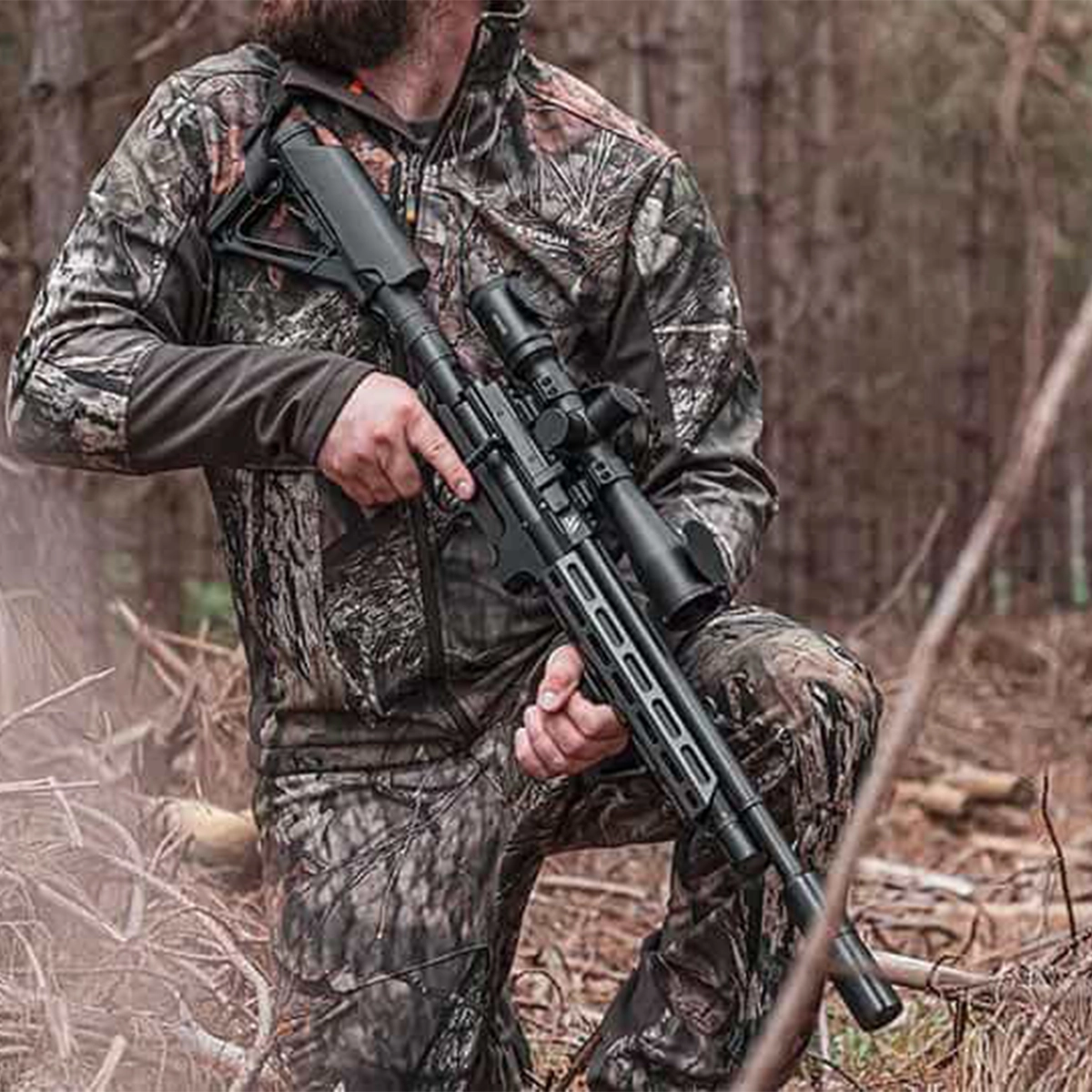 Air Arms S510 Tactical Regulated PCP Air Rifle 13 A person in camouflage kneels in the woods, holding an Air Arms S510 Tactical Regulated PCP Air Rifle fitted with a scope.