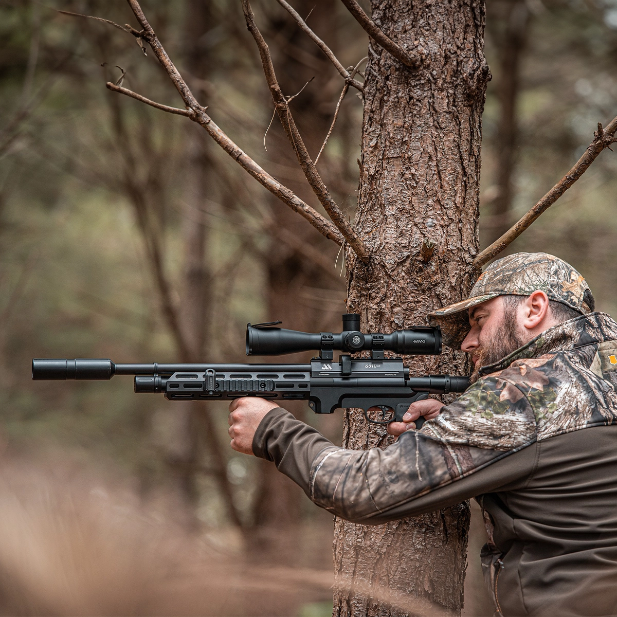 Air Arms S510 Tactical Regulated PCP Air Rifle 9 Wearing camouflage, a person aims an Air Arms S510 Tactical Regulated PCP Air Rifle while leaning against a tree in the woods.