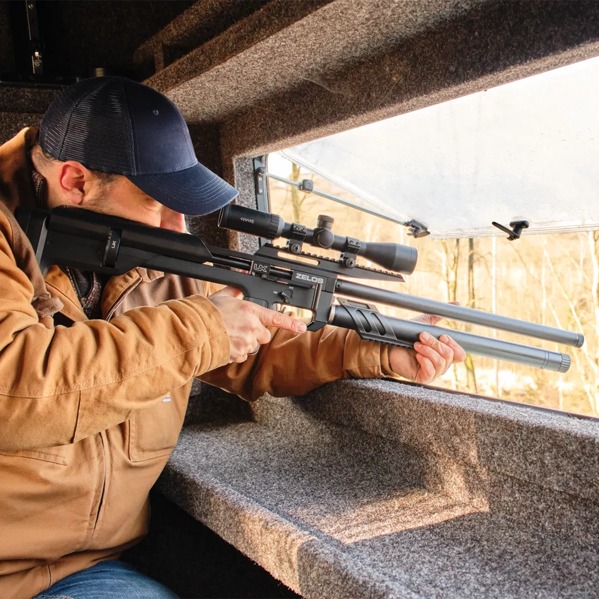 Umarex UX Zelos PCP Air Rifle 7 A person in a brown jacket and blue cap aims a scoped Umarex UX Zelos PCP Air Rifle out of a window in a camouflaged hunting blind.