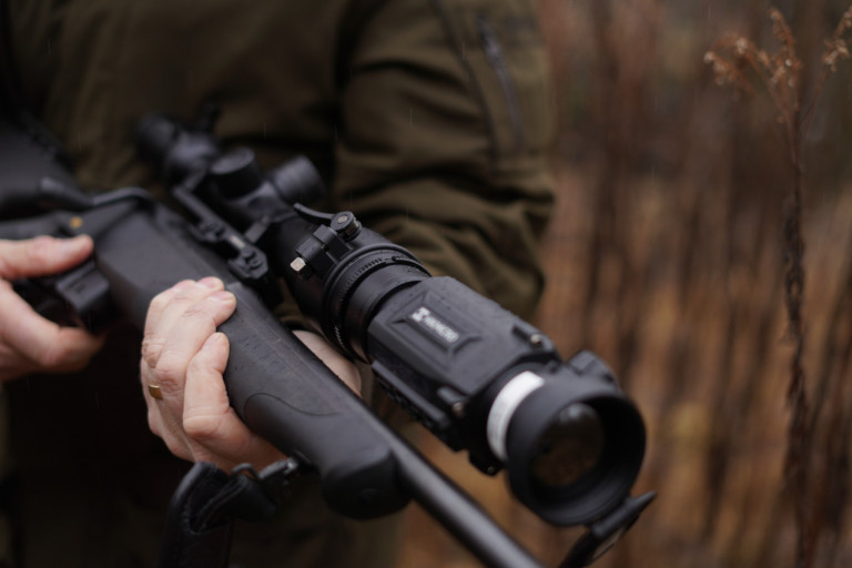 HIKMICRO Ultimate Thunder 2.0 TE19C Smart Thermal Clip-On 11 A person in a brown jacket stands outdoors with blurred brown grass behind, showcasing the HIKMICRO Ultimate Thunder 2.0 TE19C Smart Thermal Clip-On for enhanced outdoor experiences.
