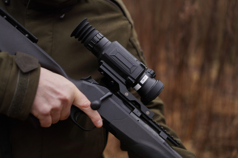 HIKMICRO Thunder 2.0 Pro TQ50 Smart Thermal Scope With Rail 9 Close-up of a person in an olive green uniform holding a HIKMICRO Thunder 2.0 Pro TQ50 Smart Thermal Scope With Rail, set against a blurred brown background.