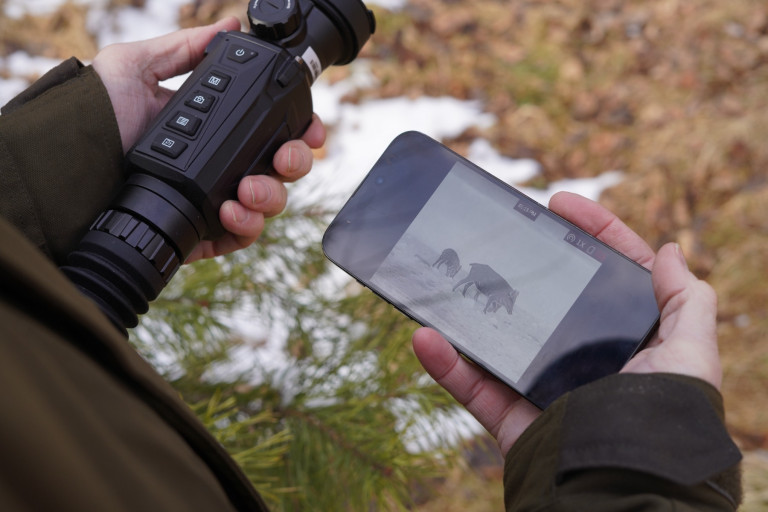 HIKMICRO Thunder 2.0 TH35 Smart Thermal Scope With Rail 5 Outdoors, someone presses a button on black headphones, with blurred brown foliage behind them—mirroring the precision of adjusting the HIKMICRO Thunder 2.0 TH35 Smart Thermal Scope With Rail in nature.